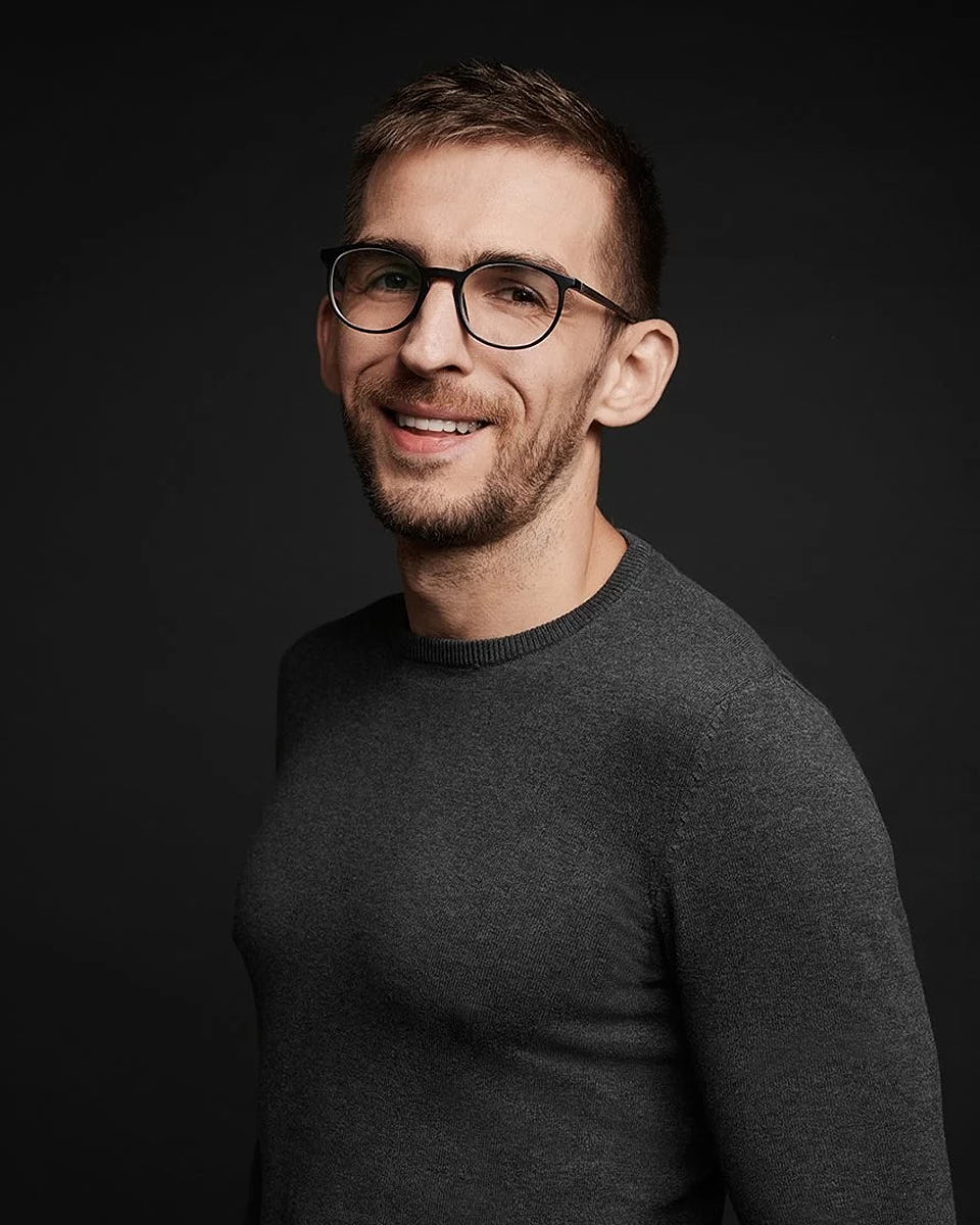Professional studio headshot of a man wearing glasses and a dark sweater against a dark background