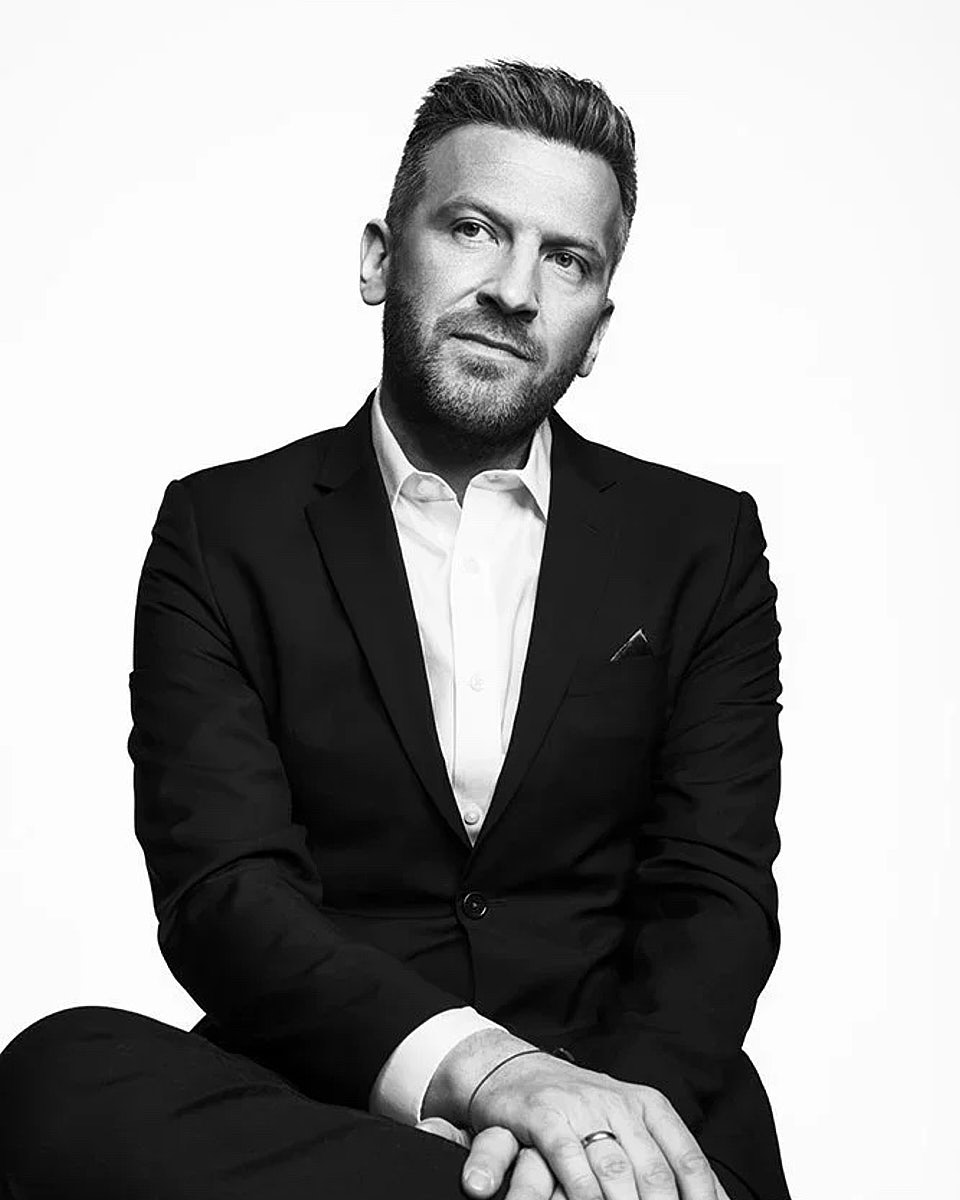 Black-and-white professional headshot of a seated man in a tuxedo-style jacket