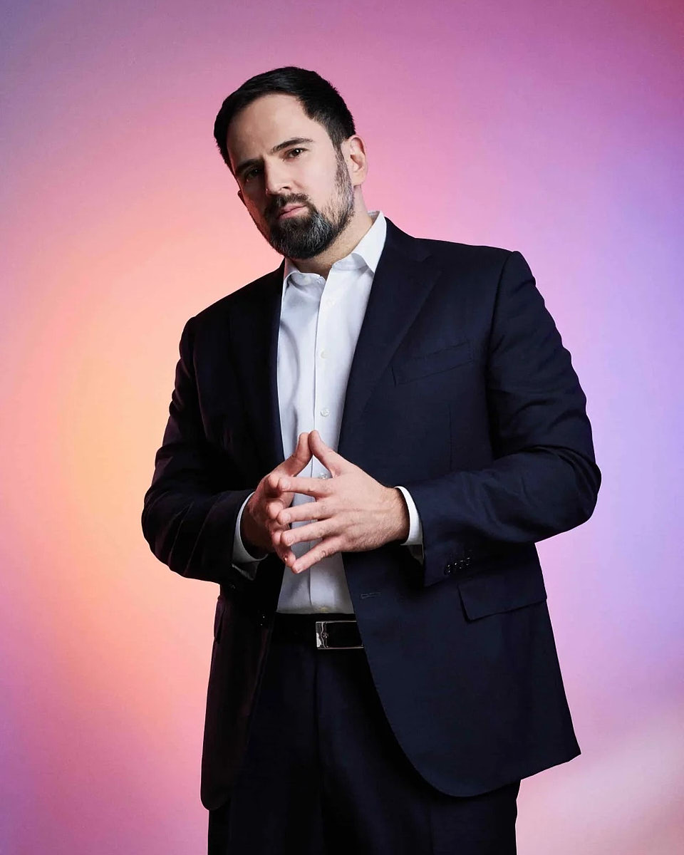 Editorial-style standing portrait of a man in a suit on a pink and purple background