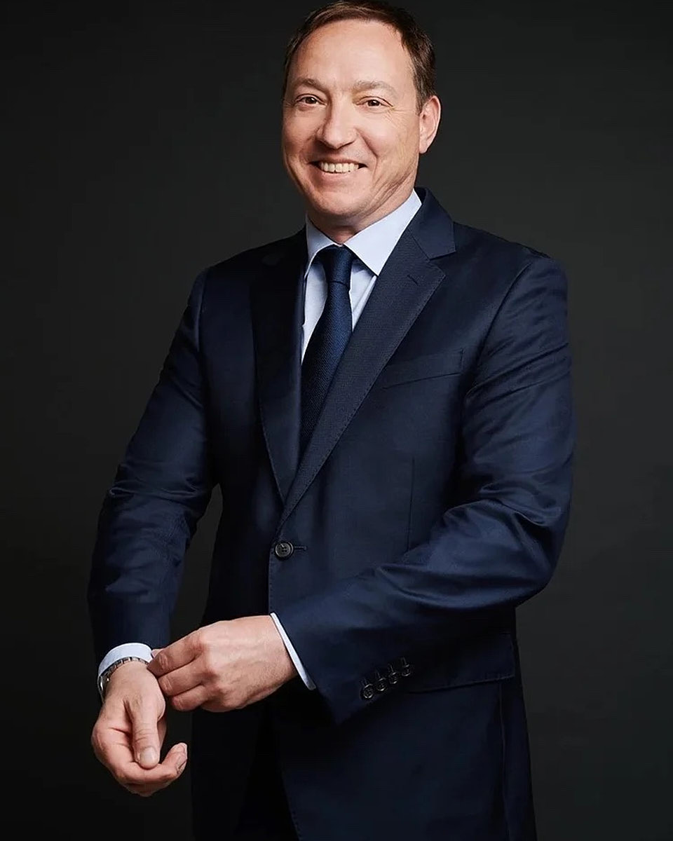 Professional headshot of a smiling man in a dark suit against a dark studio background